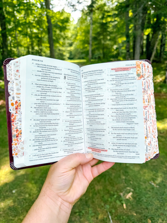 A PERSON HOLDING AN OPEN BIBLE WITH HIGHLIGHTED VERSES AND TOPICAL PRAYER TABS.  THE PERSON IS IN THE WOODS.