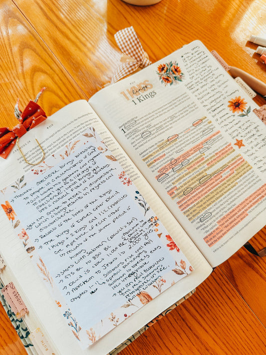 An open Bible on a wooden table with a page of notes, Bible headers, highlights, stickers, and paperclip page markers.