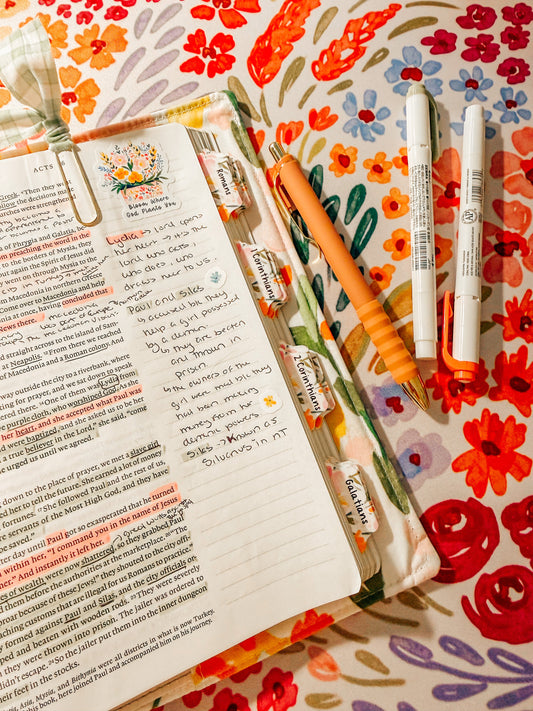 a Bible open on  a table with a floral background, pens, and highlighters.  There are notes, stickers, and highlights in the Bible.